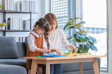 Fototapeta na wymiar Millennial Asian happy cheerful little cute preschooler daughter girl playing learning alphabets letters jigsaw toy on living room table while young teenager mother nanny helping teaching supporting