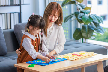 Fototapeta premium Millennial Asian happy cheerful little cute preschooler daughter girl playing learning alphabets letters jigsaw toy on living room table while young teenager mother nanny helping teaching supporting