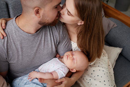 Parents Mom And Dad Kiss And Hold Newborn Sleeping Baby In Their Arms While Sitting At Home. Family Love, Close-up Portrait. Relationship Of Man And Woman Having Small Child