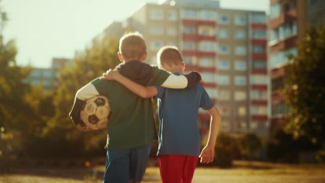 Two Young Caucasian Boys Embracing Each Other while Walking to a Neighborhood Football Pitch in Sport Clothes. Soccer Players Carrying a Ball to Play. Concept of Sports, Childhood, Friendship.
