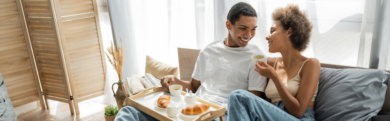 young and happy african american couple smiling near breakfast with coffee and croissants on bed, banner.