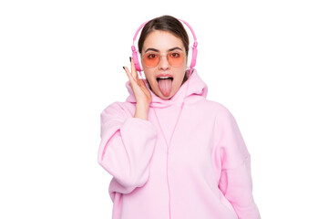 Girl in pink hoodie and glasses listening to music in headphones sticking tongue out isolated on white
