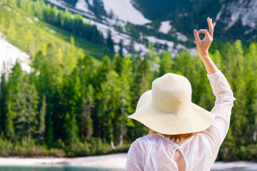 The woman in the straw hat and white dress shows the sign Ok on the background of mountains and forest. 