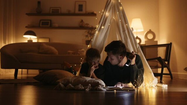 happy family spends time in home in winter evening, father and little son read book in teepee hut