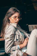 beautiful brunette girl in a sunglasses dressed in white blouse and beige coat posing in a chair. Stylish trendy fashion outlook