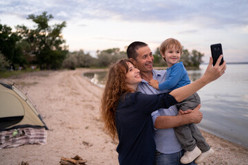 Fototapeta premium Portrait of Caucasian family with father, mother and little son selfie with lake view by smartphone while vacation together in holiday. Happy family time
