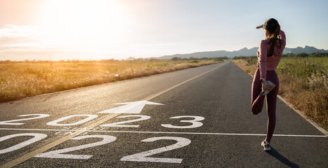 Fototapeta New year 2023 or start straight concept.word 2023 written on the asphalt road and athlete woman runner stretching leg preparing for new year at sunset.Concept of challenge or career path and change. obraz