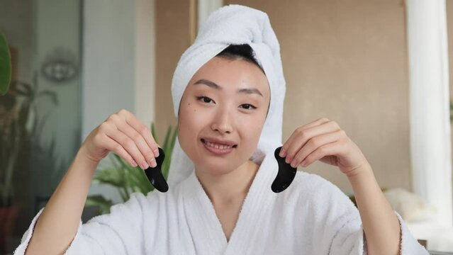 Smiling asian woman in towel on head and bathrobe sitting at table and holding collagen patches in hands. Young female using homemade cosmetic for beauty routine at exotic studio.