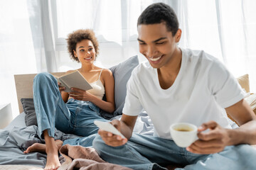 cheerful african american woman sitting on bed with book and looking at boyfriend with coffee and mobile phone on blurred foreground.