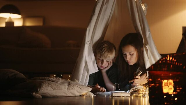 son and mom are reading magical story in fairytales book in winter evening, lying in teepee tent
