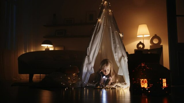 little girl reading fairytale book with lantern in night, lying in handmade tent from blankets