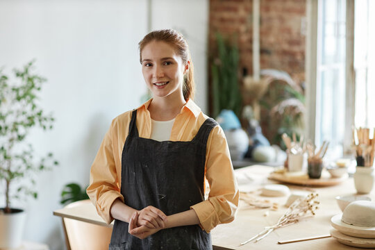 Waist up portrait of smiling young woman wearing apron while standing in pottery studio and looking at camera, copy space