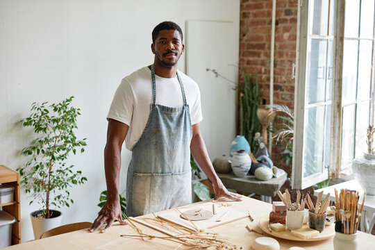 Waist up portrait of young black man wearing apron while standing in pottery studio and smiling at camera, copy space