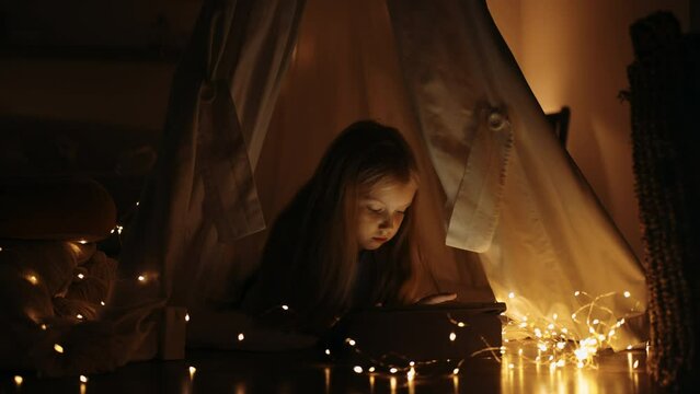 pretty chubby little girl is reading fairy tales in night, lying in tent from blanket with lantern