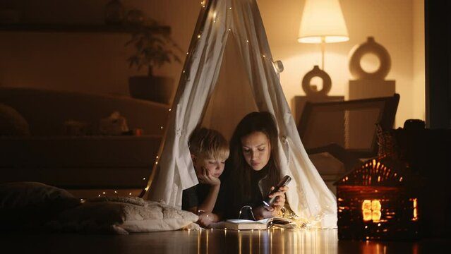 mom is reading fairytales for little son, woman and child are lying in teepee tent in living room