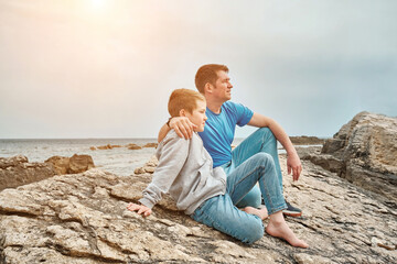 Happy family, father and son bonding, sitting on stone by the sea looking at view enjoying summer vacation. Togetherness Friendly concept