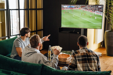 Back view of male friends watching football match and gesturing while sitting in front of the TV screen