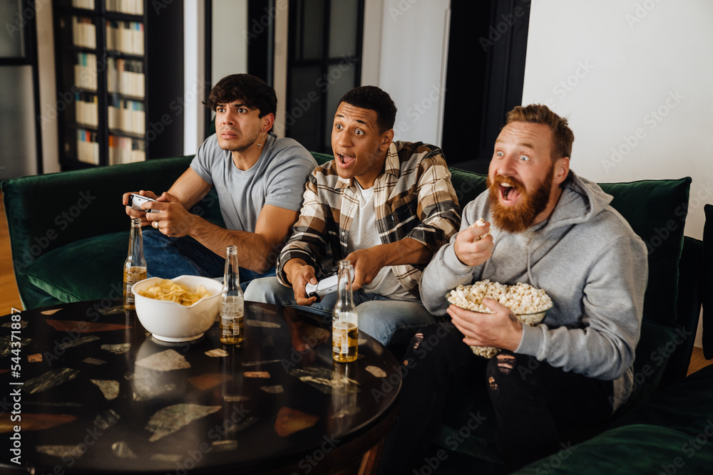 Wall mural Three excited male friends playing video games and eating popcorn in living room