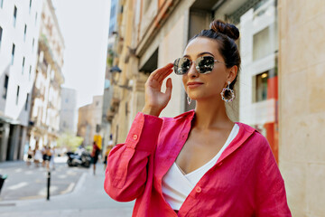 Lovely cute charming woman with dark hair wearing sunglasses and pink shirt is touching her glasses and spend time outdoor in warm summer evening 