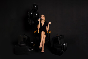 Fototapeta na wymiar Excited woman kissing credit card while sitting with balloons and shopping bags isolated over black background