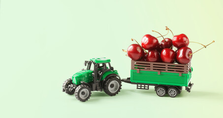 A toy tractor transports red fresh cherries in a trailer. Light green background. The concept of agricultural work, harvesting and delivery of crops. Toy world