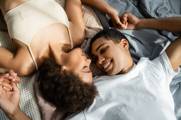 Obraz na płótnie Canvas top view of happy african american couple holding hands while lying on bed with closed eyes.