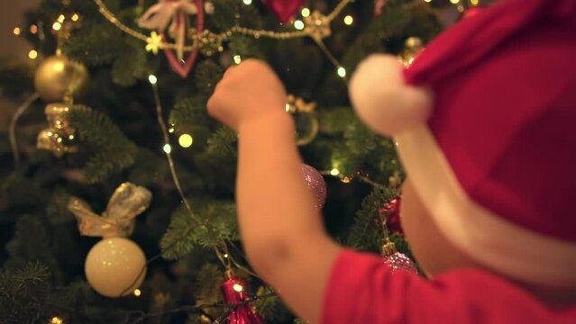 back side view little toddler kid hands santa hat trying hang shiny decoration ball Christmas tree. baby child decorating Christmas tree first time concept waiting winter holidays celebrating new year