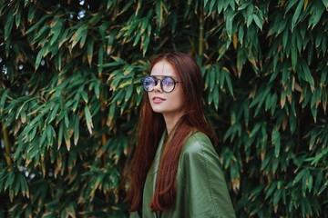 A woman in a stylish trendy green raincoat and sunglasses walks down the street near a park with bamboo