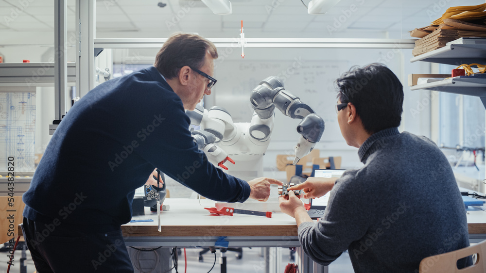 Poster Adult Caucasian Engineer Changes Position of Robotic Arm with Controller and Having a Discussion with Young Asian Developer. Startup Lab and Innovative Technologies in Robotics Concept.