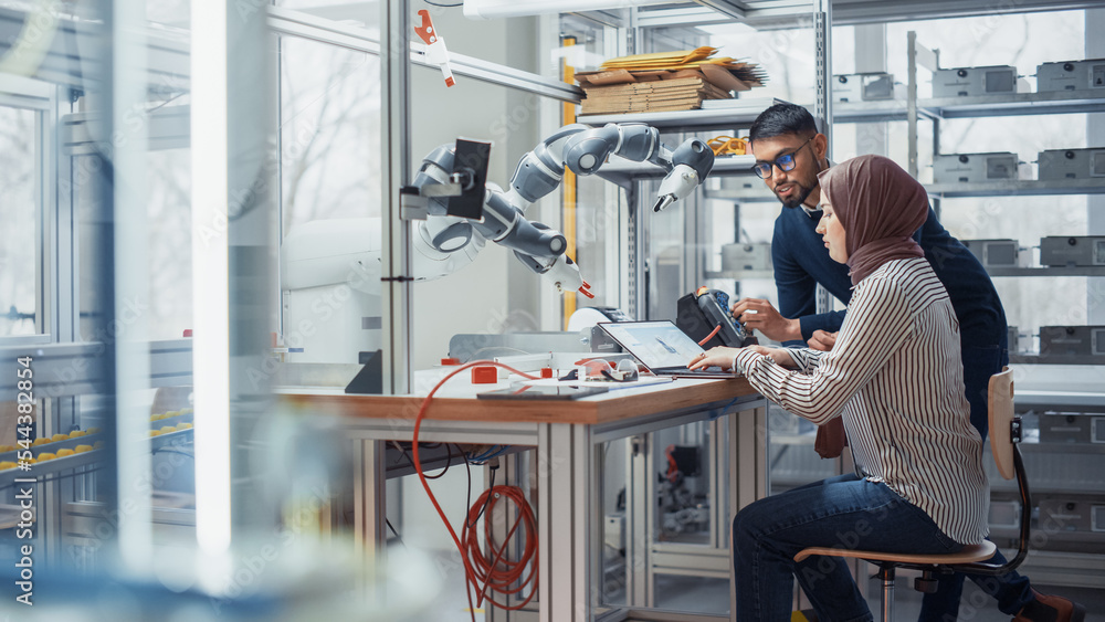 Aufkleber Black Male Engineer Raises Robotic Arm with Controller While Female Developer in Hijab Writes Code on Laptop. People Create Innovative Application. High Tech and Robotics Industries Concept.