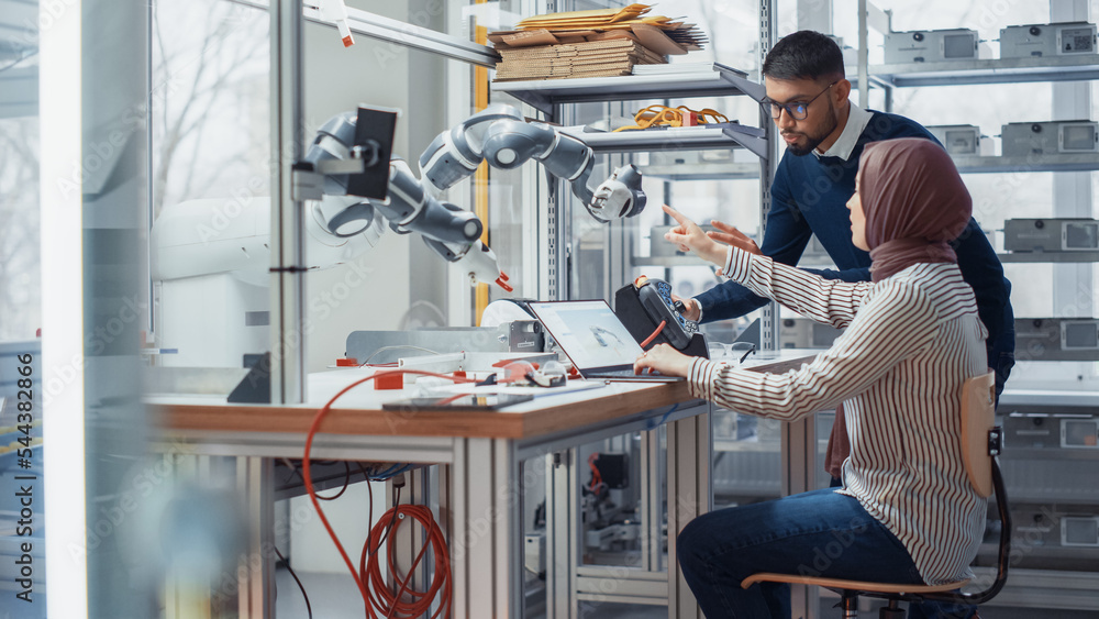 Aufkleber Professional Young Developer in Hijab Coding on Laptop While Her Black Colleague Manipulates Robotic Arm During Factory Workshop. Innovative Technologies in Automation And Robotics Concept.