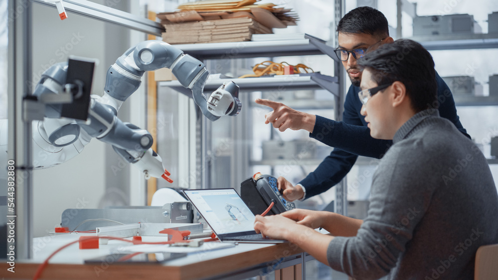 Poster Black Male Engineer Shares His Ideas With Asian Developer While Controlling Robotic Arm During Factory Workshop. People Designing Automated Robotic Systems in Startup Company Concept.