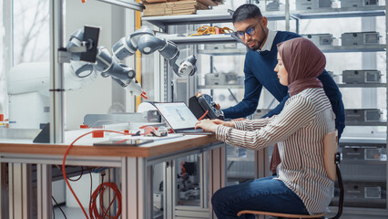 Professional Young Developer in Hijab Coding on Laptop While Her Black Colleague Manipulates Robotic Arm During Factory Workshop. Innovative Technologies And Robotics Concept.