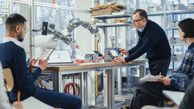 Chief Male Engineer Manipulates Robotic Arm and Talks with Young Computer Specialists in Factory Meeting Room, They Try to Find Problem Solutions. Developing and Robotics Industry Concept.