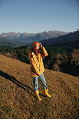 Woman walking full-length on a hill in the autumn and looking at the mountains in a yellow raincoat and jeans happy trip in the sun sunset hiking, freedom lifestyle 