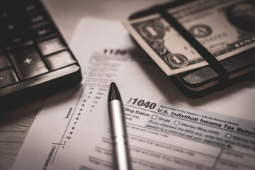 U.S. individual and corporation income tax return close-up next to a calculator, dollars, money, a pen and a notepad on a wooden table. Retro old style photo.