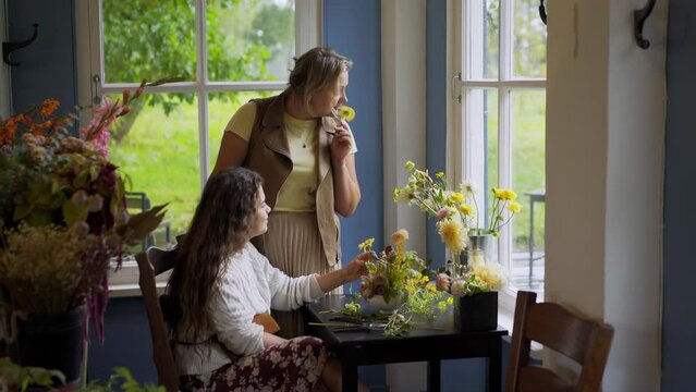 Two woman florist working together, takling and making amazing bouquet with bright fresh flowers at small table near window at arrangement masterclass
