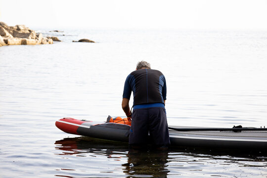 Senior Man Getting Ready To Paddle Surf On The Beach. Male Over  50 Aging Youthfully, Playing And Doing Sport At Early Morning. Older Adult Adventurer Having Fun And Enjoying. Copy Space.