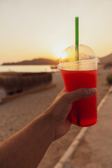 Detail of an unrecognizable man's hand holding a glass of slushy drink in the foreground at sunset on the beach.