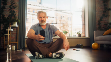 Portrait of a Strong Athletic Fit Senior Man Using Smartphone in Between Workouts During Morning Exercises at Home in Sunny Apartment. Healthy Lifestyle, Fitness, Recreation, Wellbeing and Retirement.