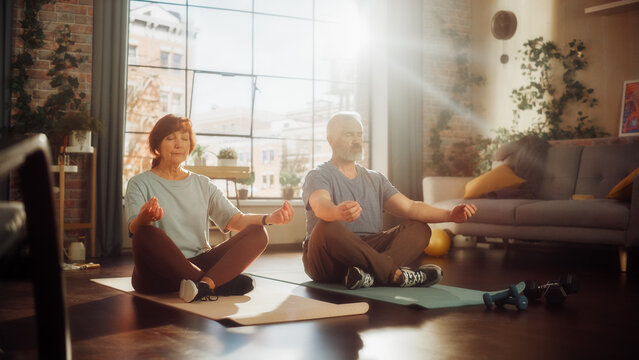 Senior Couple Meditating Together After Morning Exercises at Home in Sunny Living Room. Middle Aged Man and Woman Rest After Workout. Healthy Lifestyle, Fitness, Recreation, Wellbeing and Retirement.