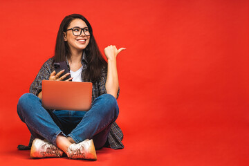 Business concept. Portrait of happy smiling brunette woman in casual sitting on floor in lotus pose and holding laptop isolated over red background. Using mobile phone.