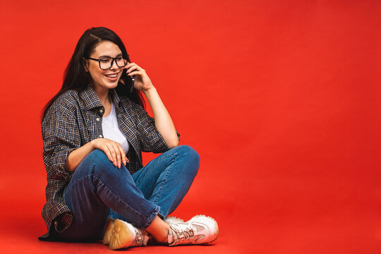 Business concept. Portrait of happy smiling brunette woman in casual sitting on floor in lotus pose and using mobile phone isolated over red background.