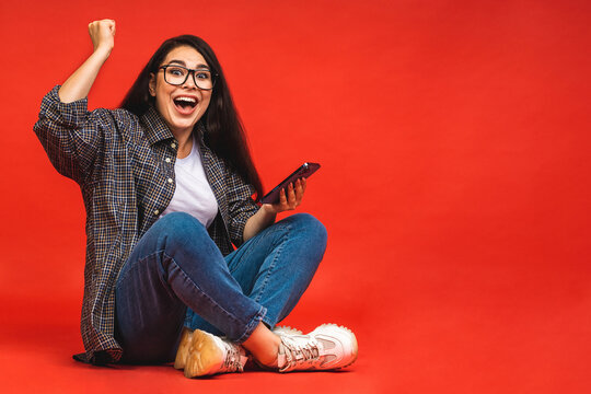Business concept. Portrait of happy brunette woman in casual sitting on floor in lotus pose and using mobile phone isolated over red background.