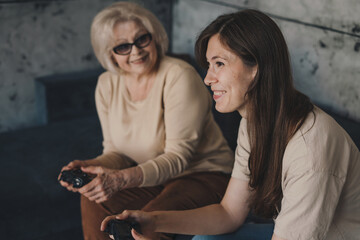 Cheerful grandmother and daughter playing videogames holding gamepad controllers sitting on couch at home. Enjoying virtual battle. Home entertainment concept.