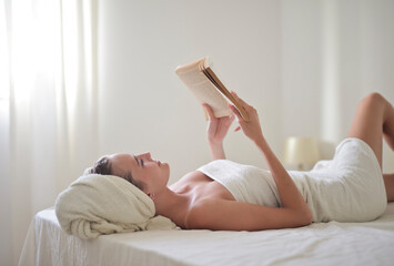 young woman lying on the bed with a towel on her head reads a book