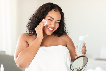 Attractive black body positive woman using facial toner and cotton pad for moisturising skin,...