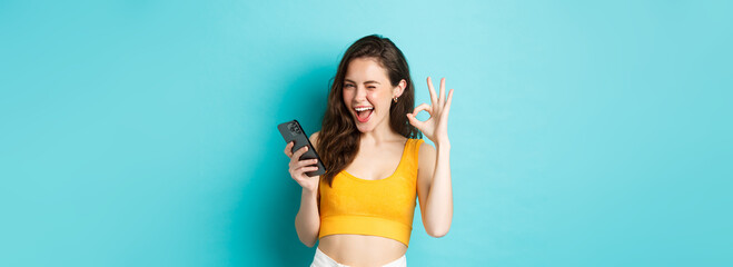 Technology and lifestyle concept. Cheerful brunette female model say yes, winking and showing okay...