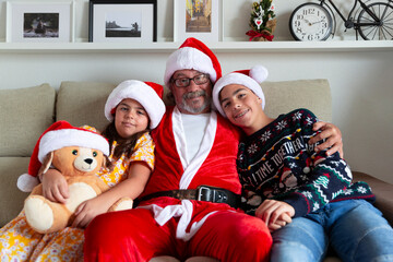 Christmas at home. Family portrait of grandfather dressed as Santa Claus hugging his grandchildren.