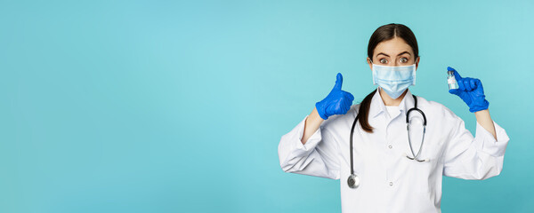 Portrait of smiling doctor, medical personel in face medical mask and rubber gloves, showing thumbs up and coronavirus, omicron vaccine, standing over blue background - obrazy, fototapety, plakaty
